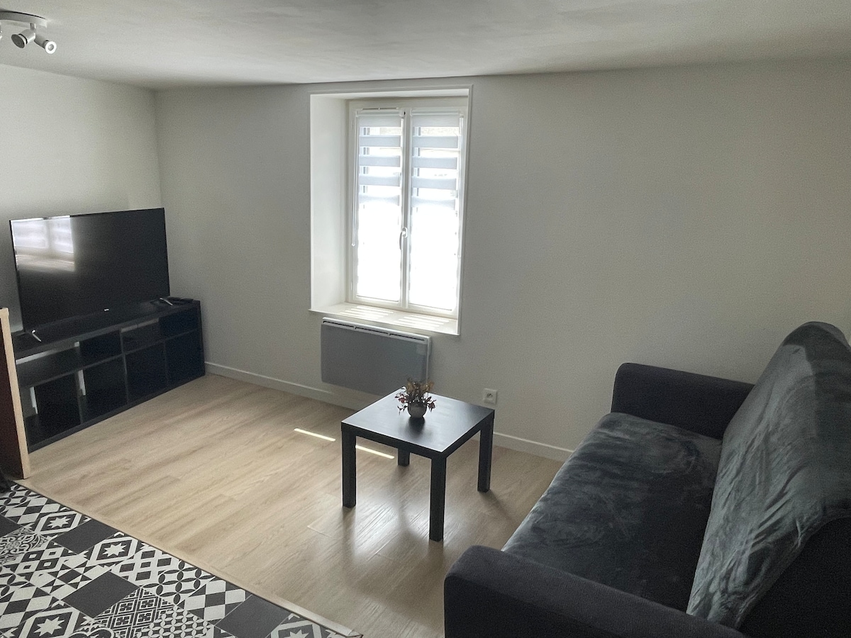 A bright and airy living area features a dark gray sofa positioned beside a small square table. A television is mounted across from the seating, and a window with partially open blinds allows natural light to fill the space.