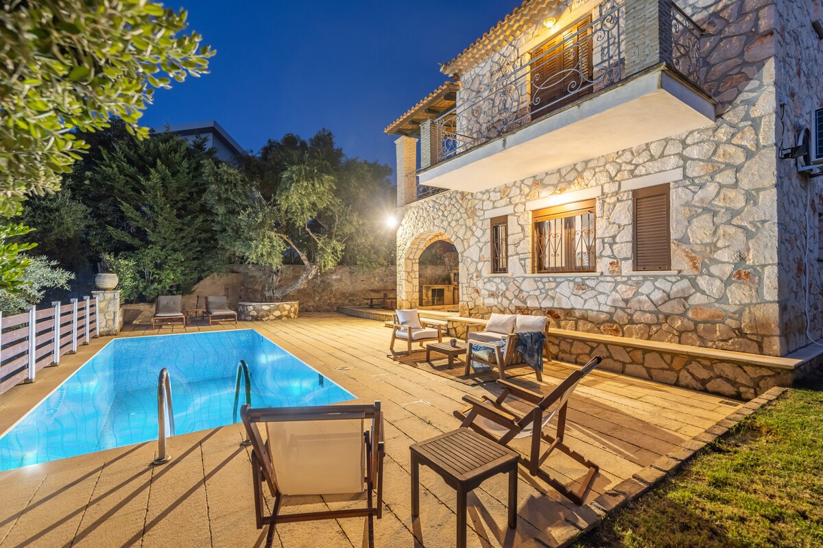 An outdoor area showcases a stone villa illuminated by warm lighting at dusk. A rectangular pool reflects the surrounding greenery, complemented by several lounge chairs positioned nearby. A spacious patio area extends from the villa, providing a comfortable space for relaxation and enjoyment.