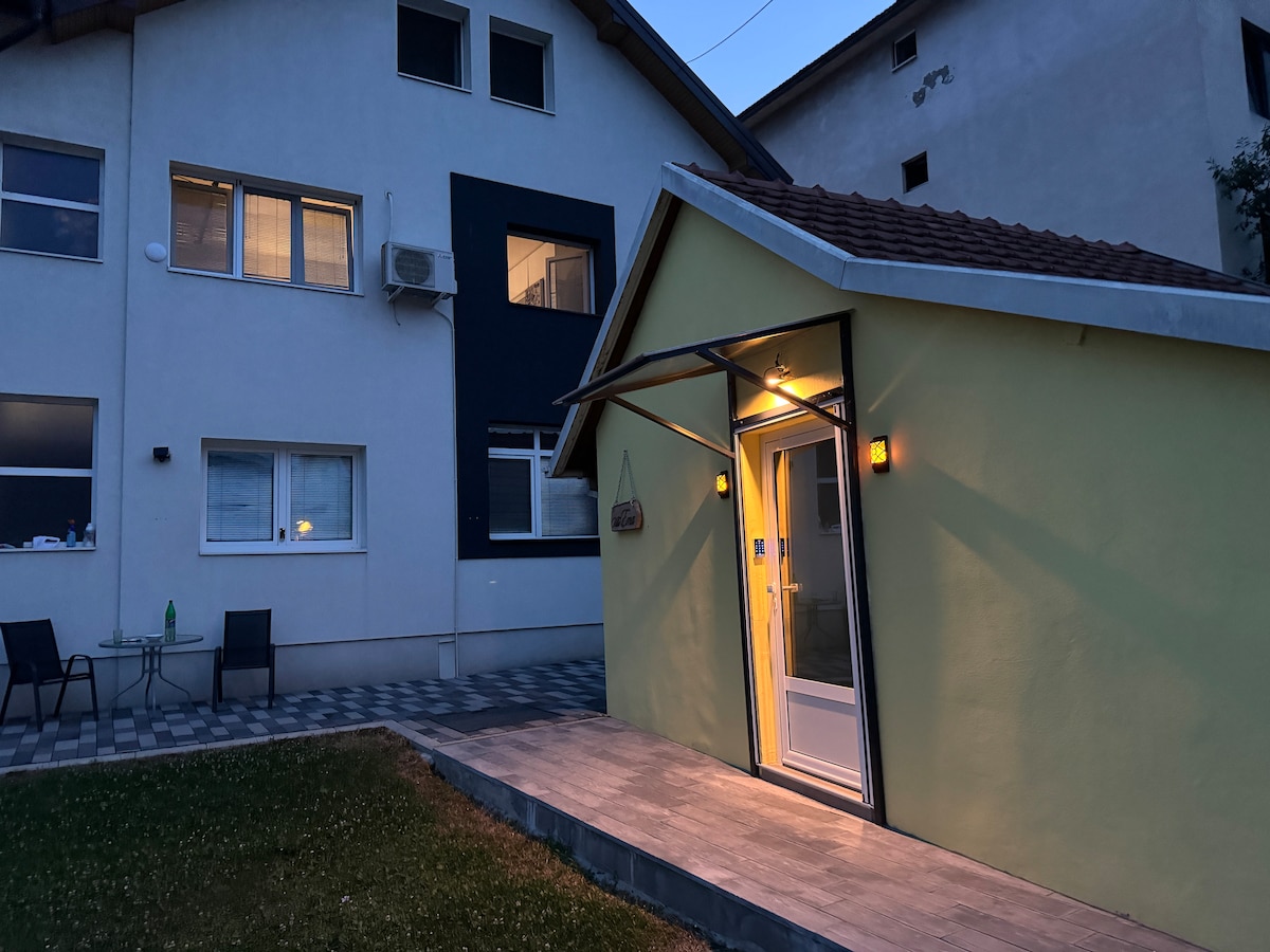 An inviting exterior view of a small yellow house is illuminated by soft lighting. A welcoming entrance is depicted, with a patio area and neatly arranged tiles. The surrounding yard features green grass, and the main building in the background showcases multiple windows.