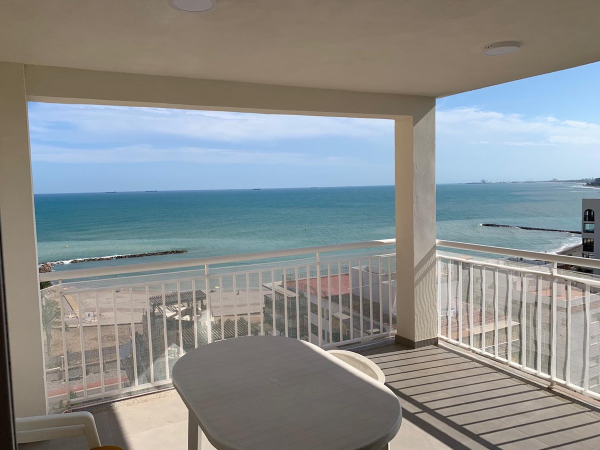 A spacious balcony offers unobstructed views of the sea, showcasing vibrant blue waters and distant ships. A simple table and chairs provide seating for relaxation, with a protective railing ensuring safety while enjoying the coastal scenery.