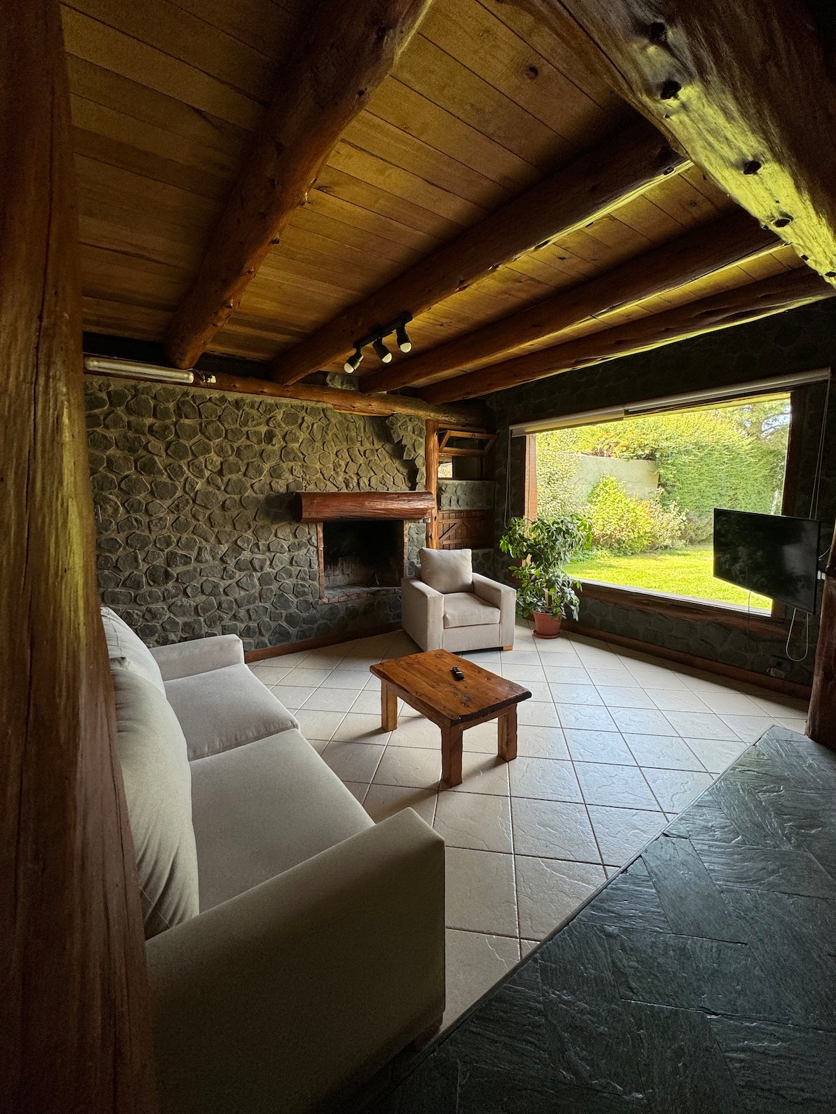 A cozy living area is highlighted by an elegant stone wall and wooden beams. Two light-colored sofas face a small wooden table, while a flat-screen TV is mounted nearby. Natural light floods the space through a large window that frames a view of greenery outside.