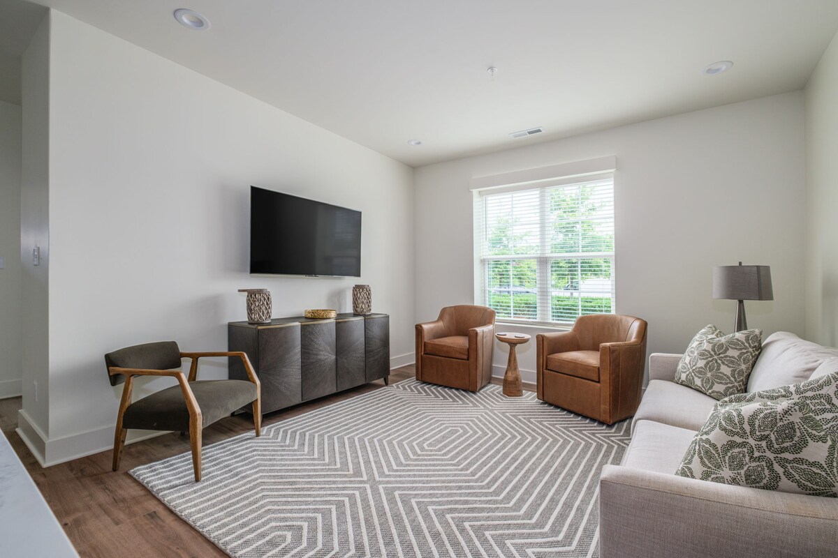 A spacious living area features a comfortable sofa, two brown armchairs, and a textured area rug. A modern television is mounted on the wall above a stylish cabinet. Natural light streams through a large window with sheer coverings, creating a welcoming atmosphere.