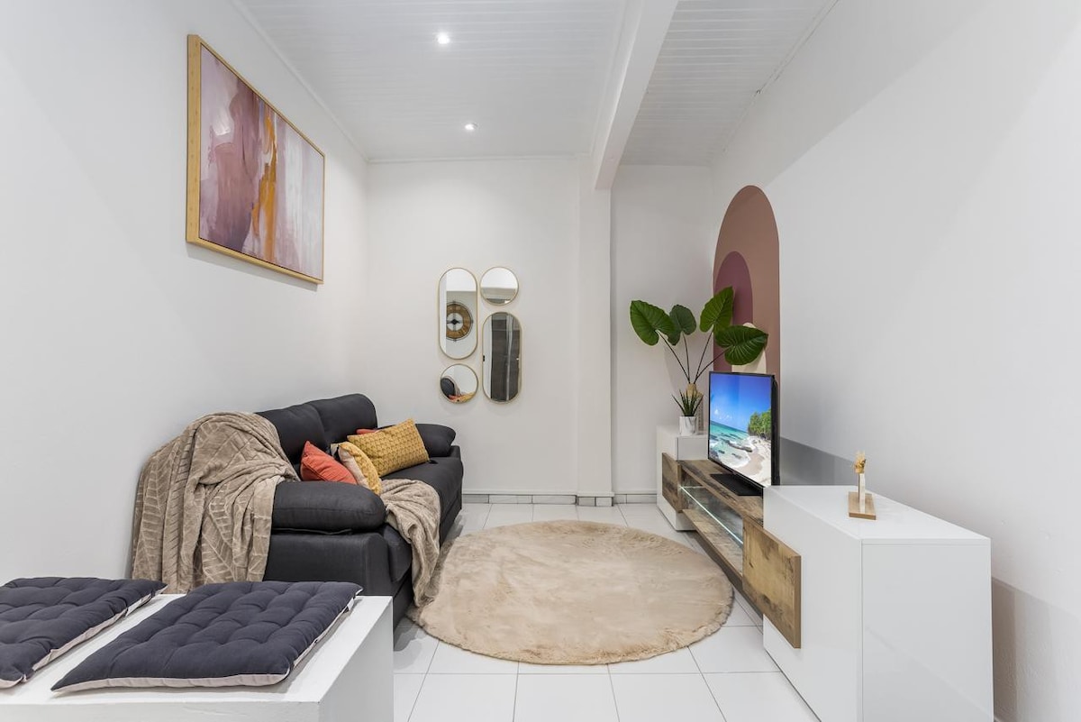 A living area features a comfortable dark sofa adorned with orange and grey cushions. A soft beige rug lies on a tiled floor, while a modern TV stand showcases a flat-screen television. Mirrors in various shapes reflect light, and a potted plant adds a touch of greenery.