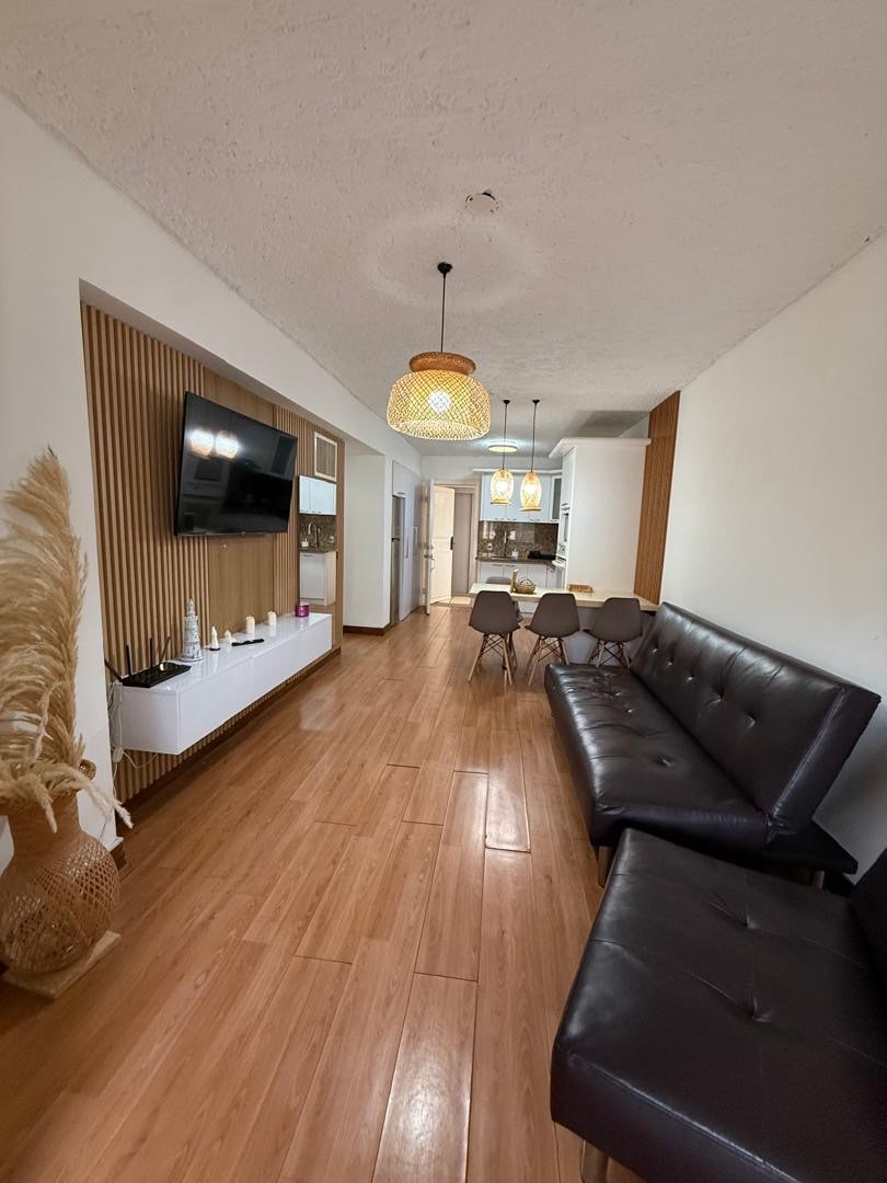 A stylish living area features a sleek dark sofa and a wall-mounted television, complemented by natural wood accents. Three pendant lights hang above a dining table surrounded by beige chairs. Light wood flooring extends throughout the space, creating a warm ambiance.