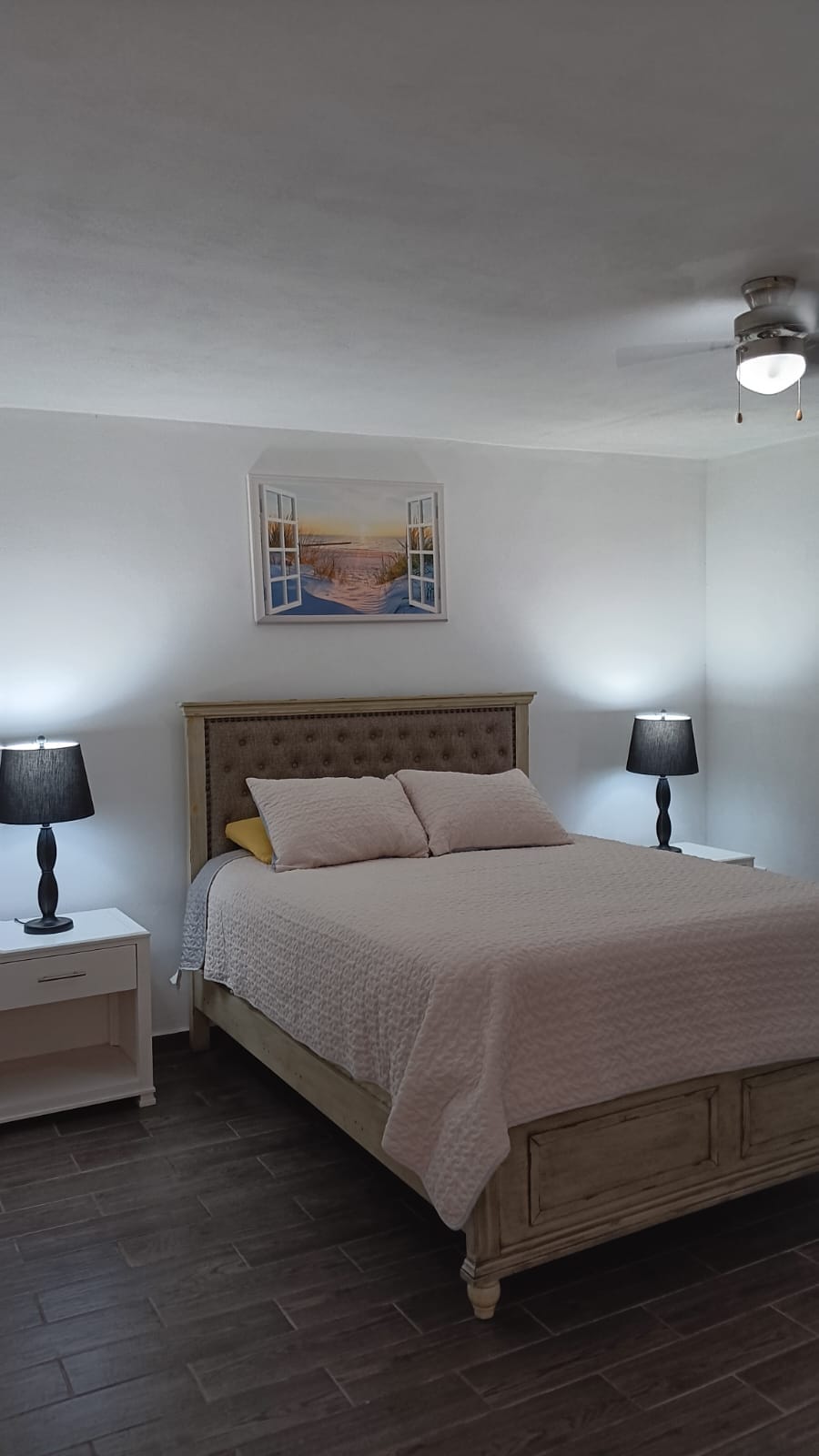 The sleeping area features a soft bed with a light-colored quilt and a tufted headboard. Two bedside lamps provide gentle illumination, and a framed artwork depicting a scenic view is mounted on the wall above. The ceiling fan offers additional air circulation.