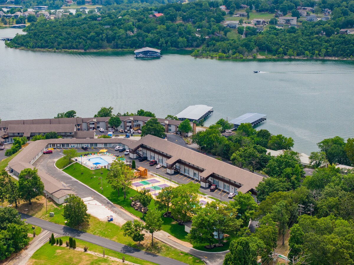 An aerial view captures the serene layout of the resort, showcasing the outdoor pool surrounded by lush greenery and amenities. The nearby lake glistens in the sunlight, with boat docks visible along the shoreline, providing access for water activities.