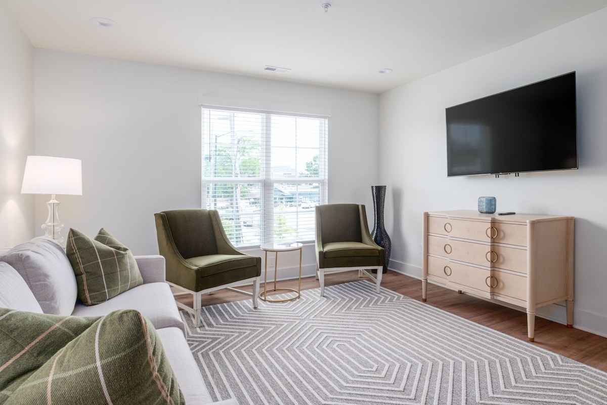 A bright living area features a comfortable sofa and two accent chairs, accompanied by a small round table. A large television is mounted on the wall, and a stylish dresser is positioned nearby. Light enters through a set of windows dressed with simple blinds.