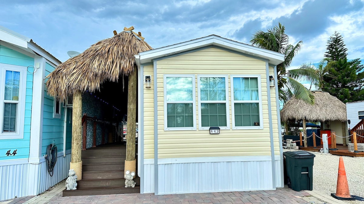 A charming seaside cottage with a light yellow exterior is presented, featuring large windows that allow natural light in. A tiki hut is visible to the left, adding to the coastal vibe. The entrance is approached by steps, enhancing accessibility.