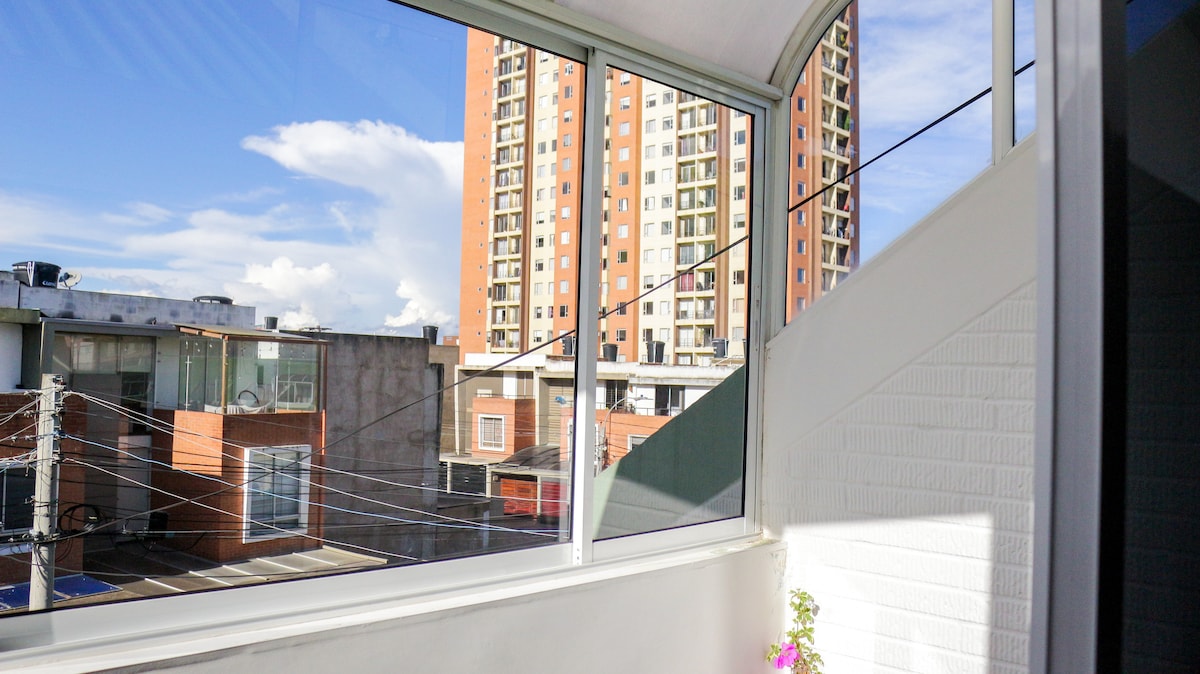 A large window offers a view of the surrounding neighborhood, showcasing nearby buildings and clear blue skies. The sleek glass design enhances natural light, while a flower pot adds a touch of color to the interior space.