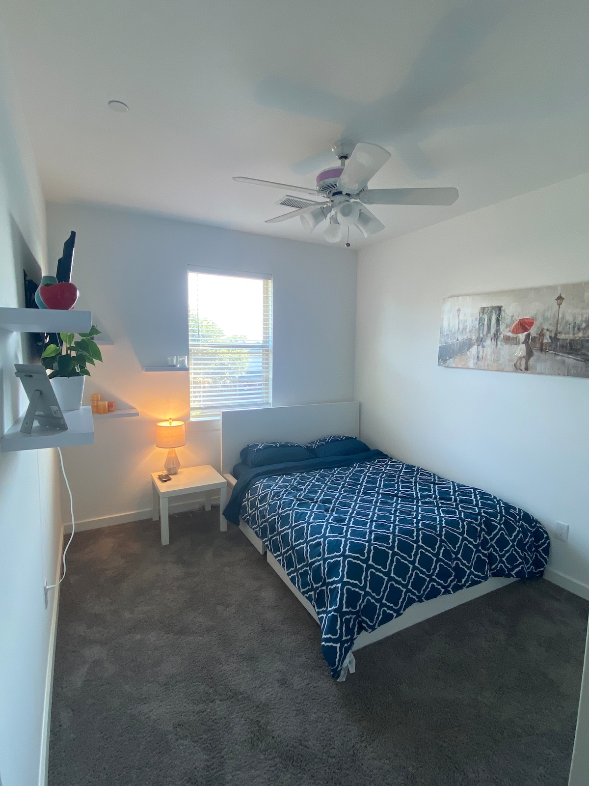 A well-lit bedroom features a spacious bed with a geometric blue-patterned blanket. A small bedside table holds a lamp, casting a warm glow. A ceiling fan is installed above, and a window with blinds allows natural light to enter, while a wall art piece adds a subtle decorative touch.