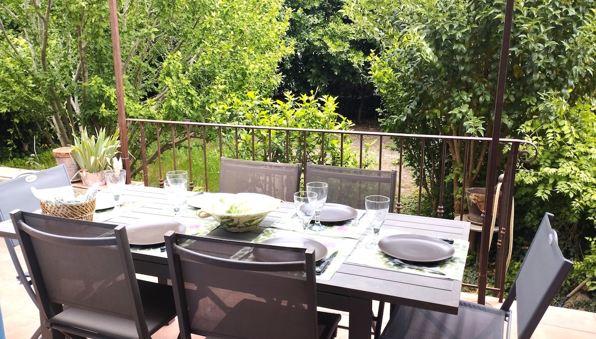 An outdoor dining area is set on a terrace, featuring a wooden table surrounded by several chairs. A tablecloth is adorned with place settings, while greenery from surrounding trees provides a serene backdrop. Sunlight filters through the leaves, creating a relaxed environment.