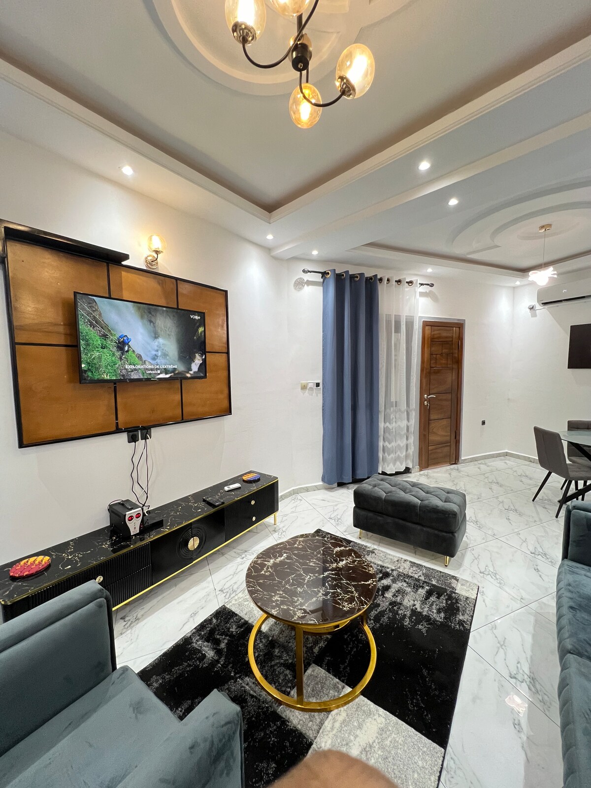 A modern living area features a stylish dark coffee table positioned on a soft black and white rug. Comfortable seating is provided by two plush armchairs in a muted blue shade. A flat-screen television is mounted on the wall, complementing the elegant decor.