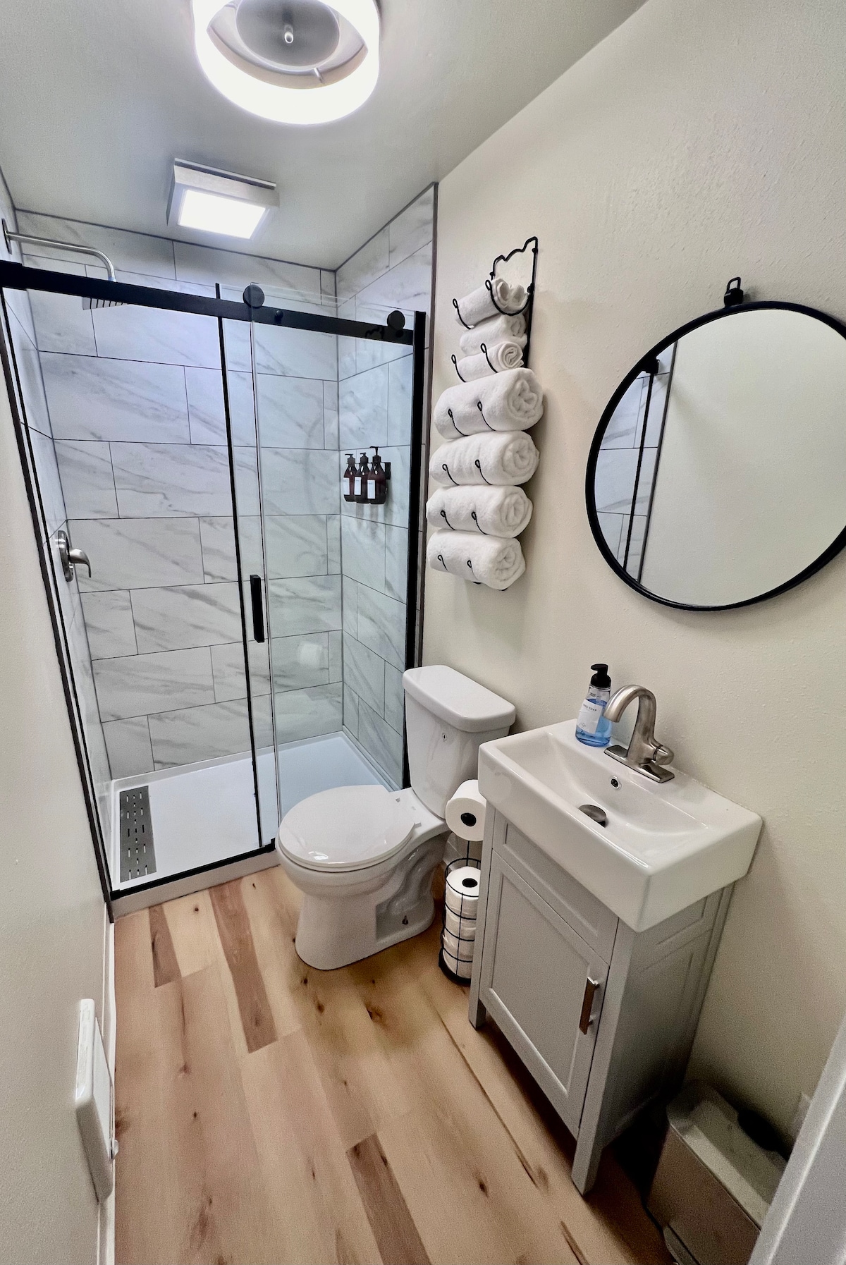 A modern bathroom features a walk-in shower enclosed with glass doors. A compact vanity with a sink is positioned next to a toilet. Fresh towels are neatly arranged on a shelf, while a round mirror reflects the light from the ceiling fixture.