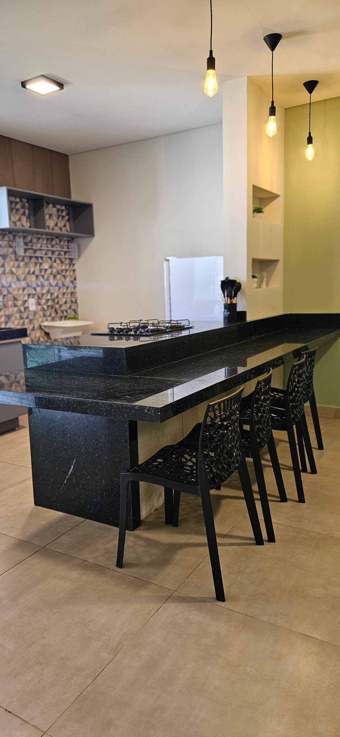 A modern kitchen is equipped with a sleek black countertop and bar seating for four, featuring stylish black chairs. The walls showcase a geometric tile design and shelves for storage. Natural light brightens the space, highlighting the white appliances in the background.