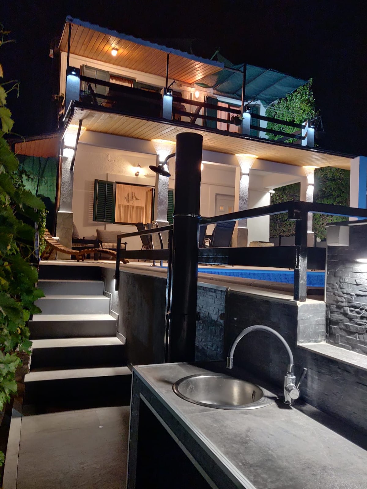 A modern outdoor area features a sleek outdoor sink and a staircase leading up to a two-story structure. The building is illuminated at night, highlighting the elegant architectural details and surrounding greenery, creating a tranquil atmosphere.