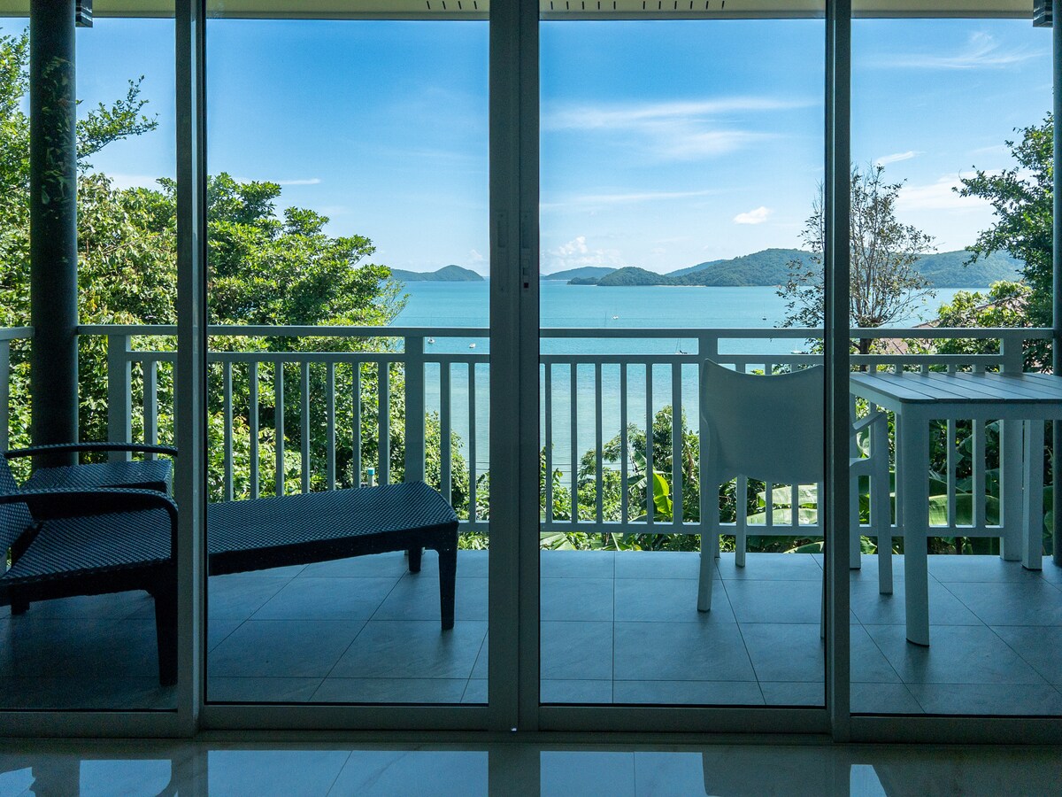 A balcony view showcases a serene sea landscape framed by lush greenery. Large glass doors open to the outdoor area furnished with a white table and chairs, contrasting against the backdrop of distant islands and a clear blue sky.