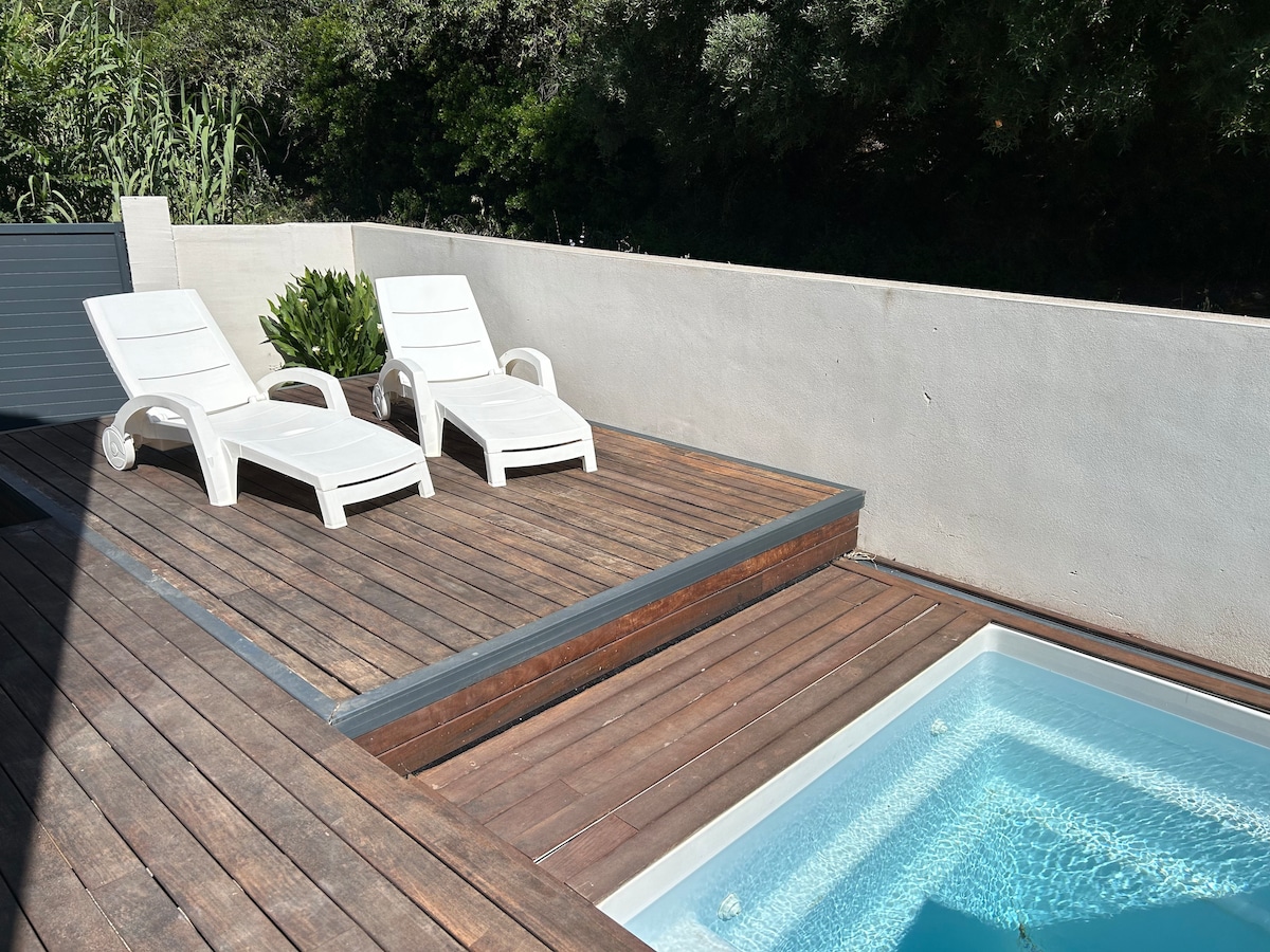 A wooden deck features two white lounge chairs positioned beside a small swimming pool. The pool has clear blue water and is surrounded by a white wall and greenery, creating a serene outdoor relaxation area.