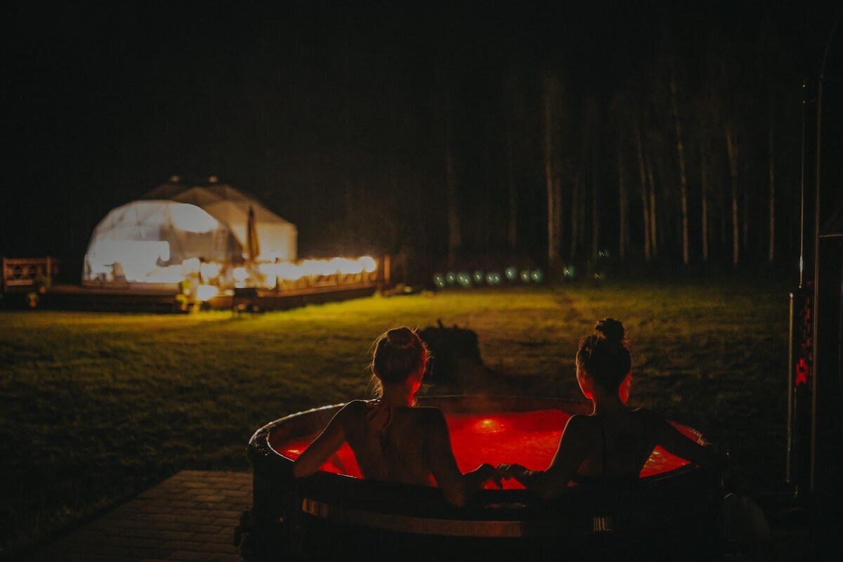 Two individuals are seated in a hot tub with softly illuminated red lights, creating a serene atmosphere at night. In the background, a dome-shaped house is visible, surrounded by trees and softly lit, enhancing the setting's peaceful ambiance.