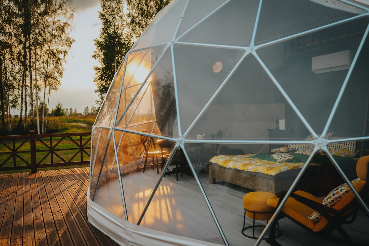 A geodesic dome house is showcased on a spacious wooden deck, surrounded by nature. Large transparent panels allow natural light to enter, highlighting the inviting interior with a cozy bed and minimalist furnishings. Tall trees frame the scene, enhancing the sense of tranquility.