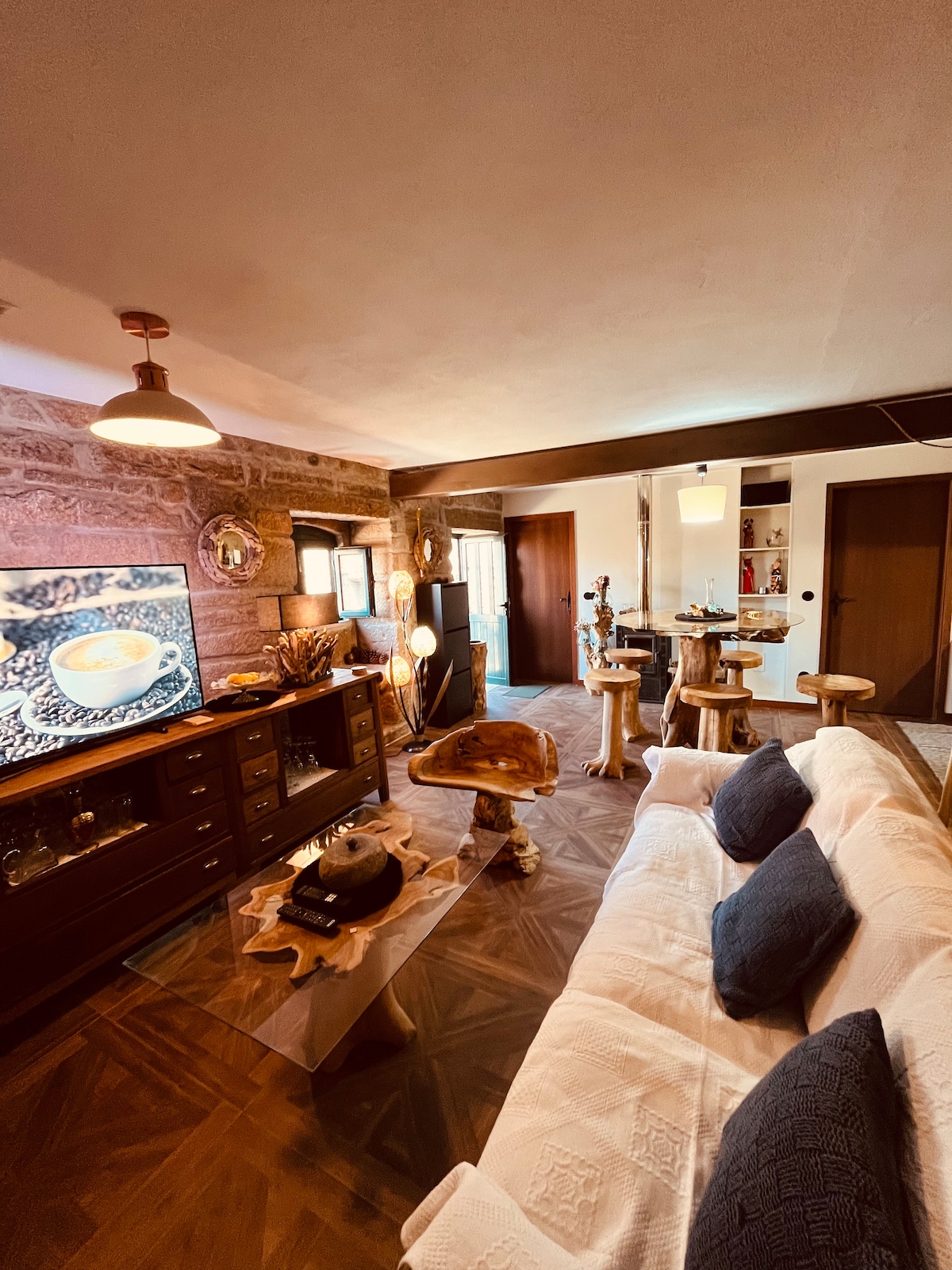 A cozy living area features a light-colored sofa adorned with blue cushions, positioned near a wooden coffee table. A television sits on a dark wooden cabinet, while unique wooden stools are arranged around a small table. Stone walls and warm lighting contribute to a welcoming atmosphere.