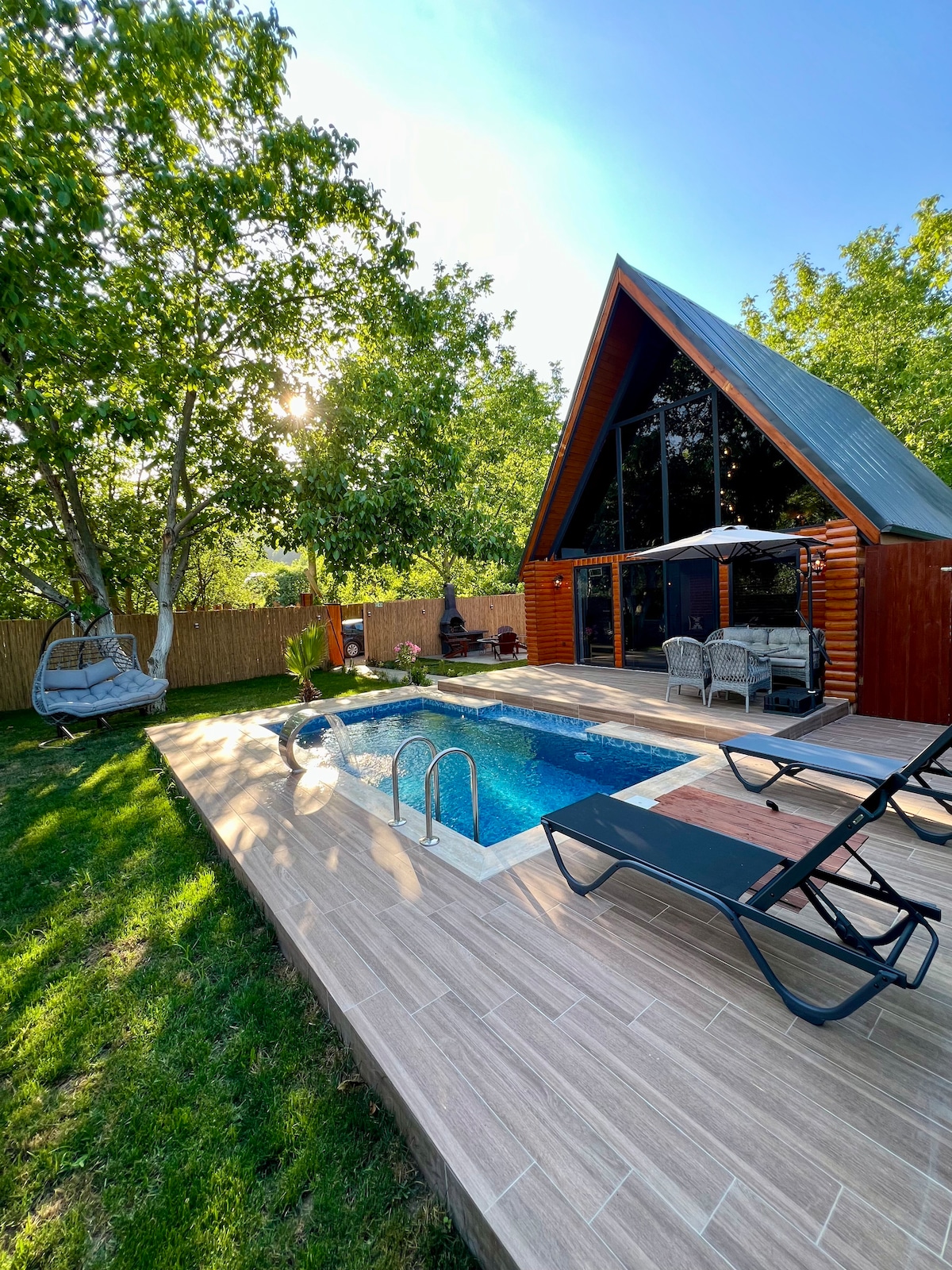 A wooden cabin features a triangular roof and large glass panels, allowing natural light to fill the interior. Surrounding greenery enhances the outdoor area, which includes a private pool, sun loungers, and a shaded seating space under an umbrella.