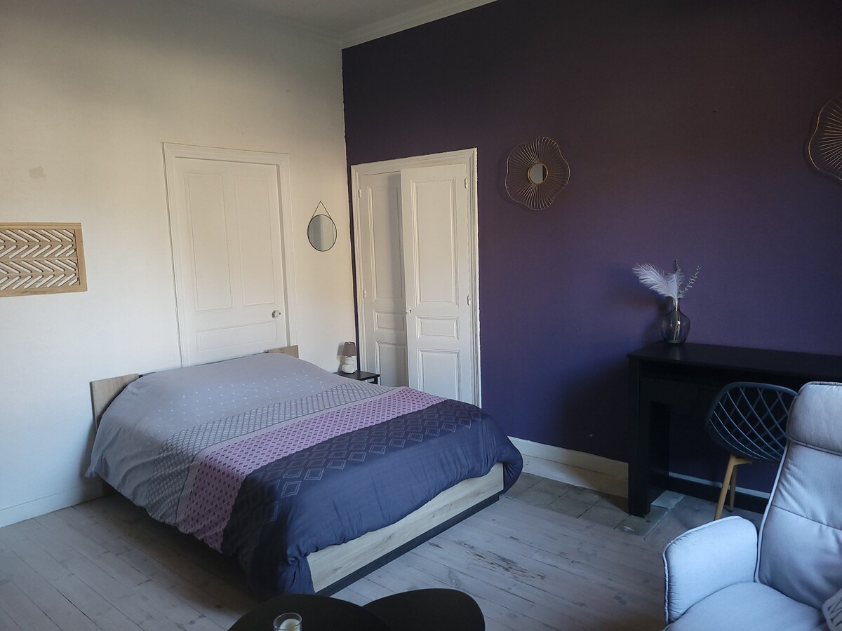 A spacious bedroom features a large bed adorned with muted tones, complemented by purple accent walls. A desk sits against one wall, with a chair nearby. Two doors lead to other rooms, while natural light illuminates the space.