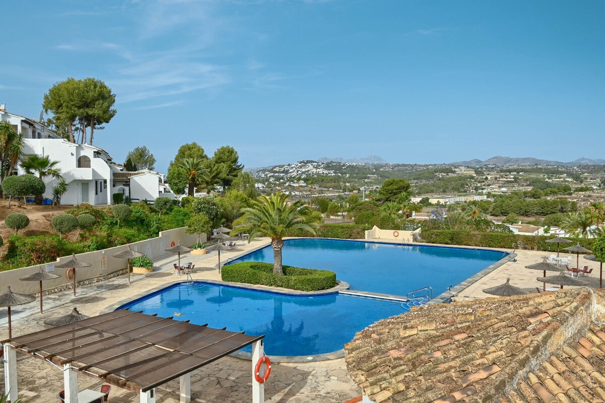 A large community pool is surrounded by landscaped greenery and palm trees, offering tranquil views of the surrounding hills. Lounge chairs are positioned around the pool area, and a shaded pergola provides seating for relaxation. Clear blue skies complete the inviting outdoor space.
