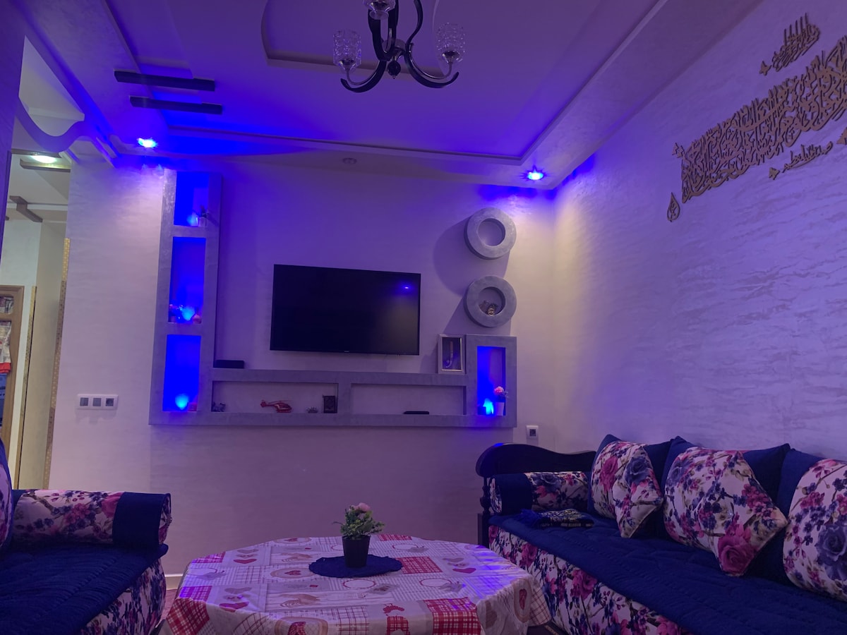 A modern living area features a floral-patterned sofa with plush cushions, arranged around a circular coffee table. Ambient blue lighting enhances the room's aesthetic. A mounted television is set against a textured wall, accompanied by decorative shelves displaying a few items.