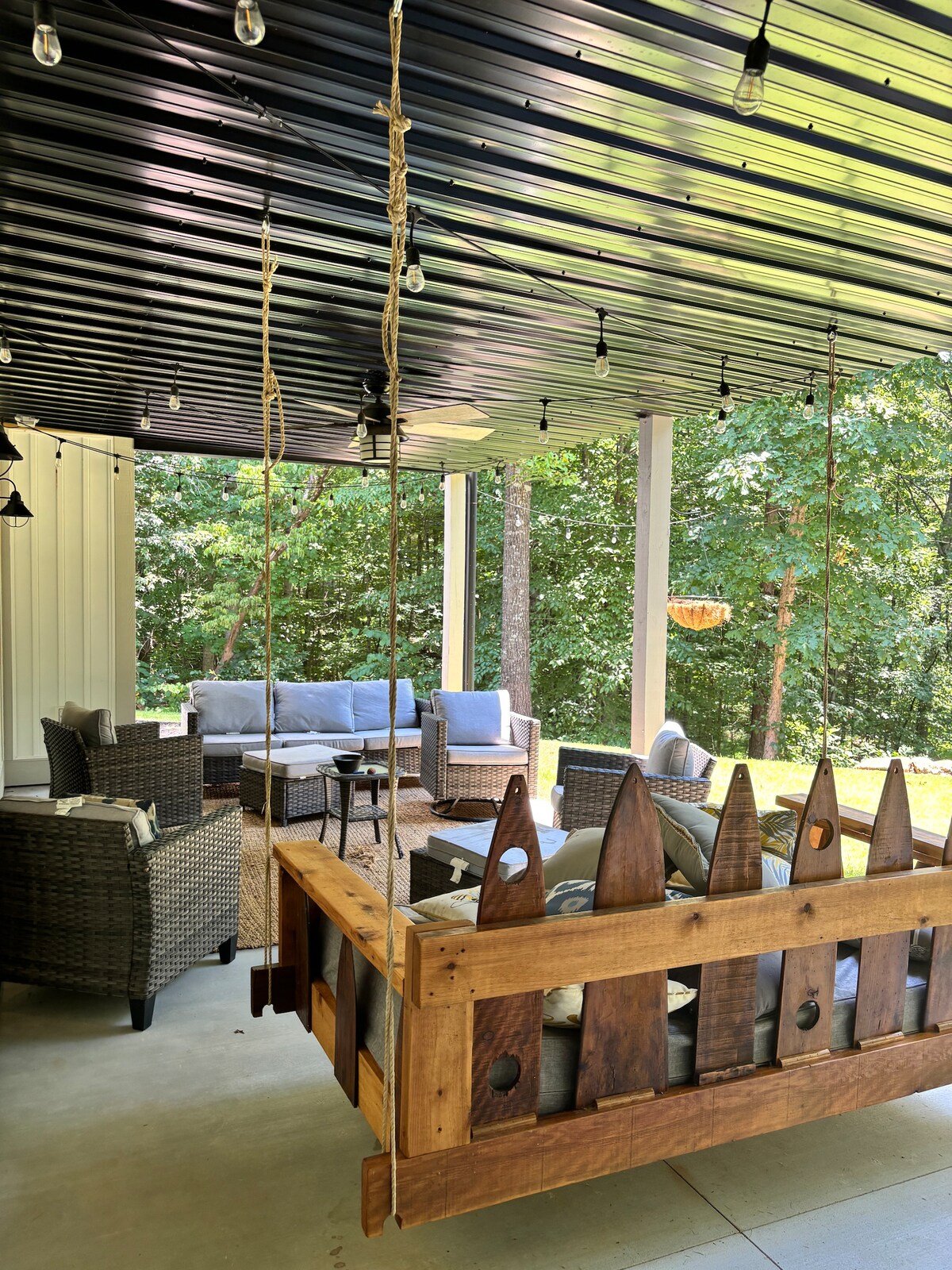 A covered outdoor area features a wooden swing surrounded by comfortable seating arrangements. Soft lighting hangs from the ceiling, illuminating the space. Greenery is visible in the background, creating a serene setting for relaxation.