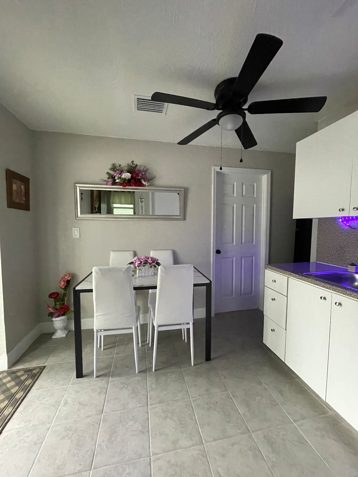 A dining area is seen featuring a glass table surrounded by four white chairs. A mirror with decorative elements is mounted on the wall. A ceiling fan is installed above, and kitchen cabinets are visible in the background with LED lighting underneath.