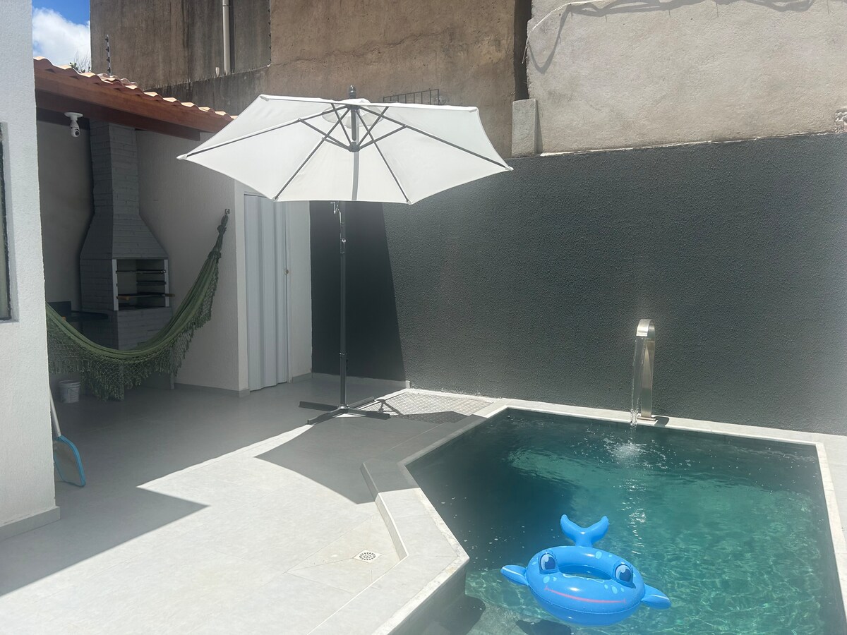 A private pool area is visible, featuring a sleek design with a shallow section and a small waterfall. A large white umbrella stands adjacent to the pool, providing shade. A blue inflatable float in the shape of a whale is placed on the water's surface.