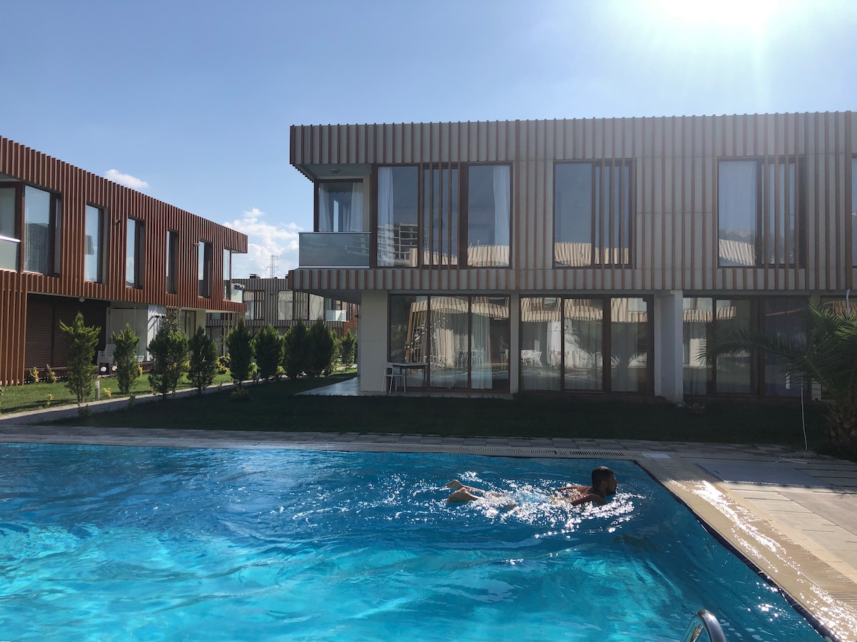 A modern building with wooden accents is situated next to a clear, inviting pool. Large glass windows reveal a well-lit interior. Lush greenery surrounds the pool area, contributing to a serene outdoor environment.