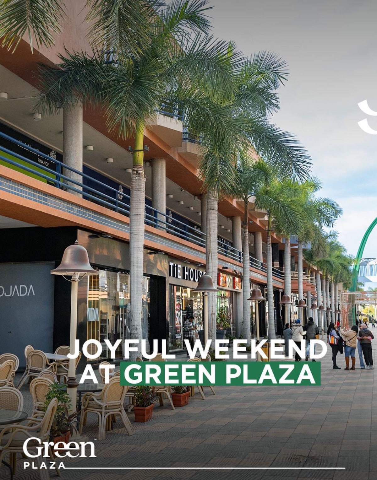 A vibrant plaza is depicted, lined with palm trees and colorful storefronts. The spacious pathway invites visitors to explore various shops and eateries. Green planters are positioned along the walkway, contributing to the lively atmosphere and providing a welcoming space for families to enjoy their time together.
