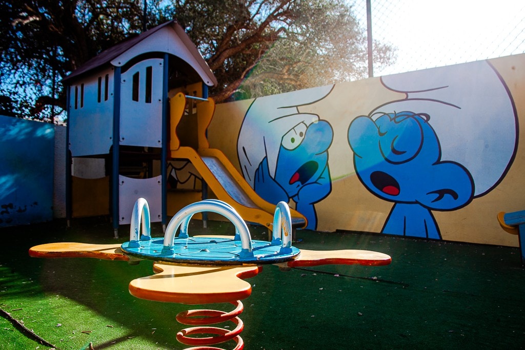 An outdoor play area features a colorful children's playground with a slide and a playhouse. A spinning toy sits in the foreground, while a vibrant mural depicting cartoon characters adds a playful touch to the space. Soft grass underfoot creates a safe environment for play.