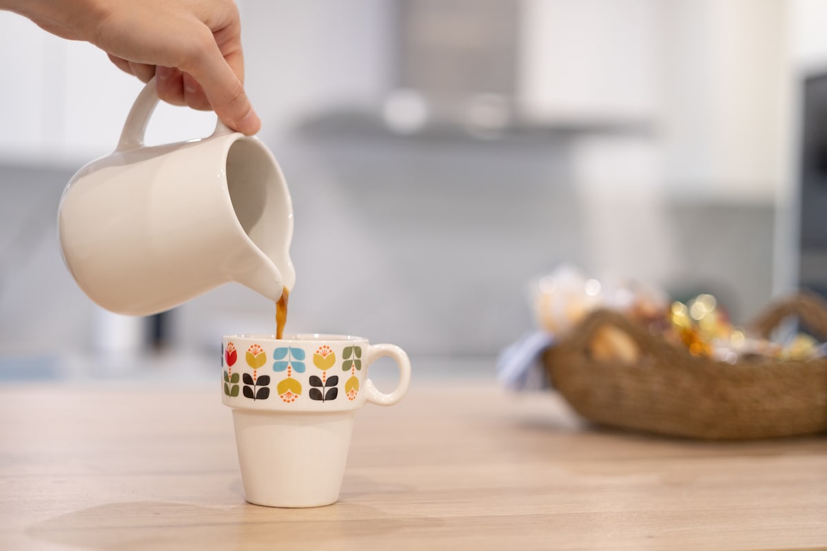 A white pitcher is tilted, pouring coffee into a decorative cup featuring colorful floral designs. The setting includes a wooden table, with a basket in the background, containing various snacks, enhancing the welcoming atmosphere of the kitchen space.