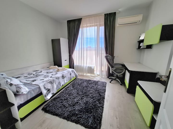 Un dormitorio luminoso con una cama individual, muebles de tonos verdes y una alfombra suave junto a la cama. Incluye un armario y un escritorio, una gran ventana con vistas y aire acondicionado para una estancia cómoda. Perfecto para trabajar o relajarse.