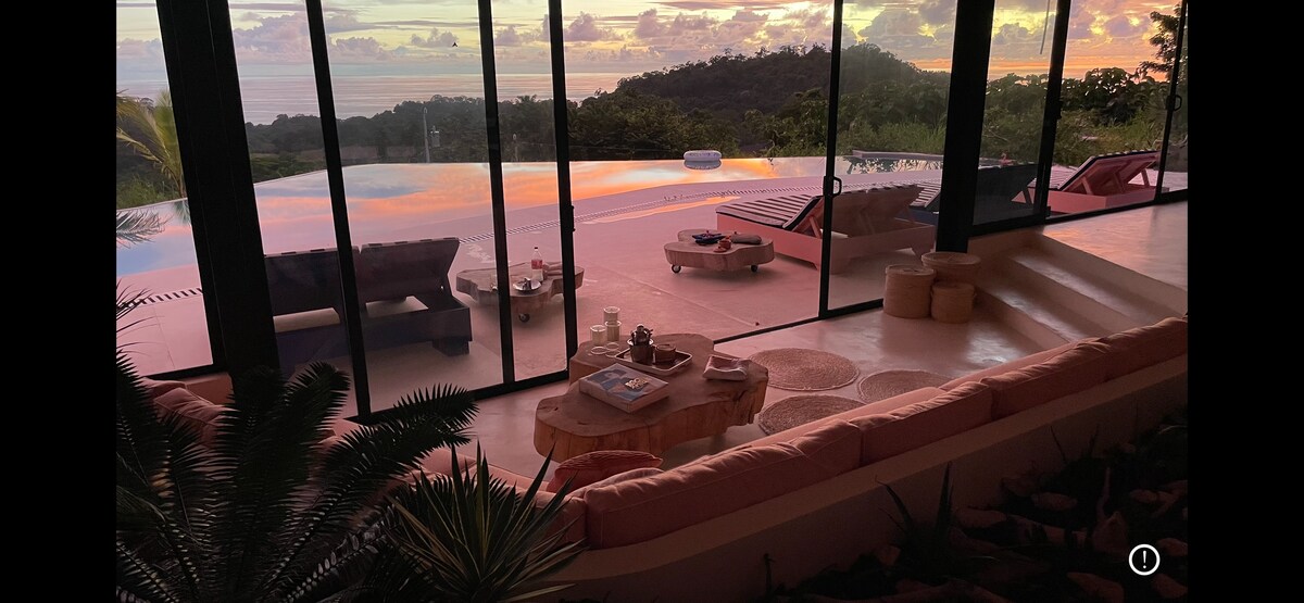 An inviting lounge area features a spacious sofa adorned with soft cushions, facing large glass doors that provide expansive views of the infinity pool and sunset over the ocean. A coffee table sits in the center, complemented by decorative items and a low seating arrangement nearby.