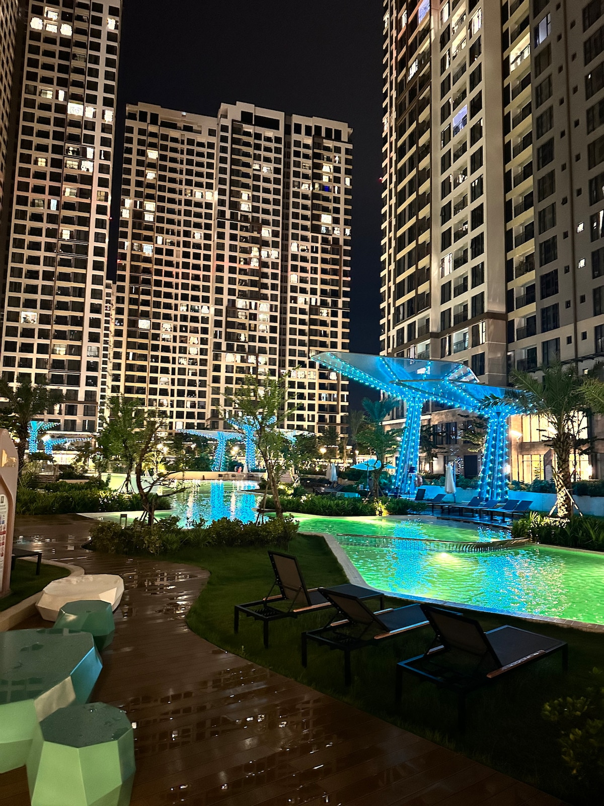 A tranquil evening scene showcases a spacious pool area surrounded by sleek buildings. Soft blue lights illuminate the water, enhancing the lush greenery and modern loungers. Pathways wind through landscaped gardens, inviting relaxation and enjoyment of the outdoor environment.