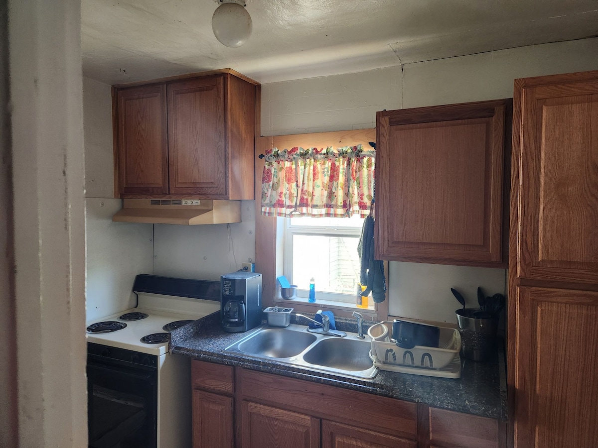 A functional kitchen with wooden cabinetry and a floral curtain over the window. The countertop features a double sink, a microwave, and multiple cooking appliances. The tiled backsplash and the array of kitchen utensils add to the practicality of the space.