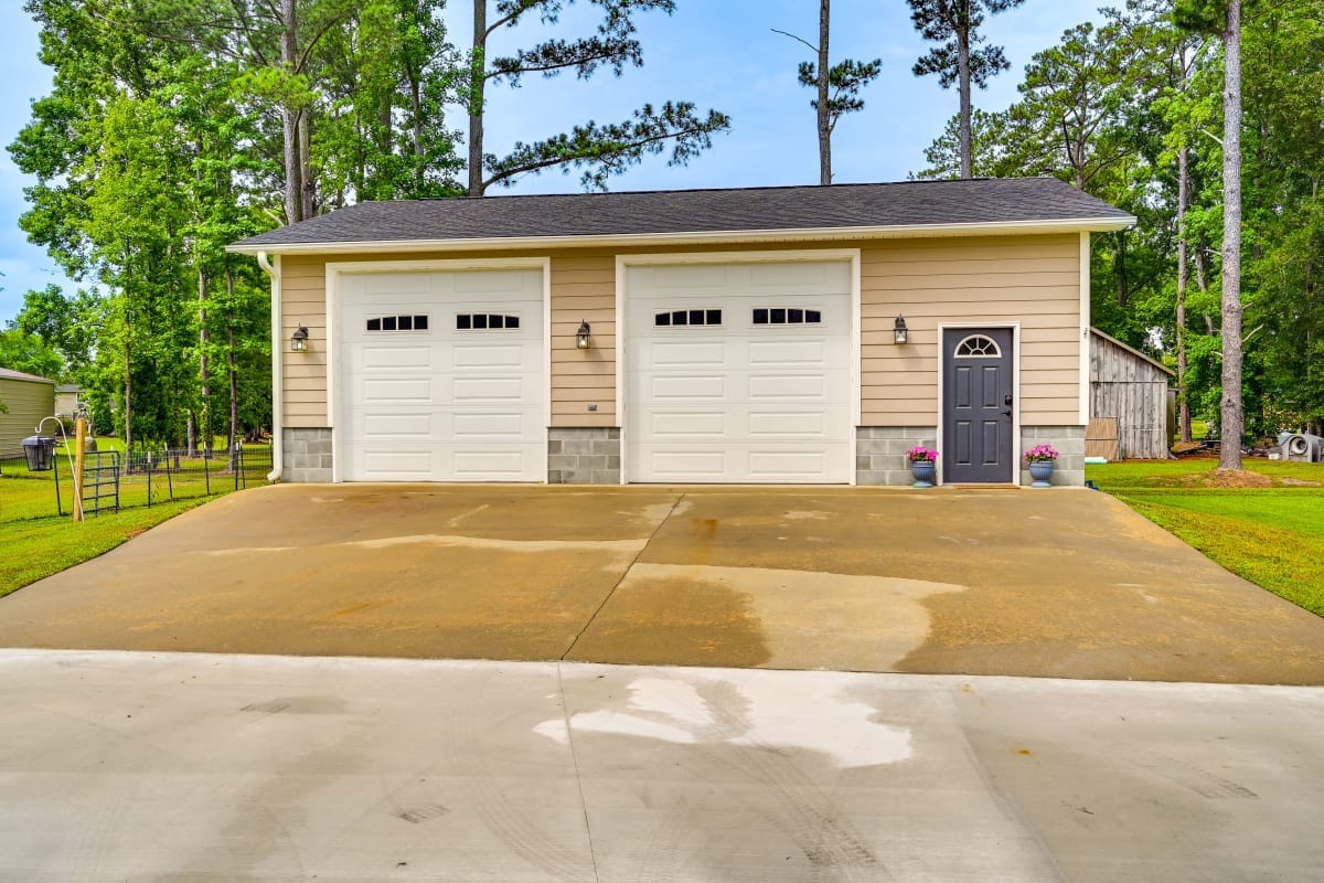 A two-car garage features white doors with decorative windows, set against a neutral exterior. The driveway is wide and leads to the entrance, bordered by green landscaping. The setting is surrounded by tall trees, providing a sense of privacy.