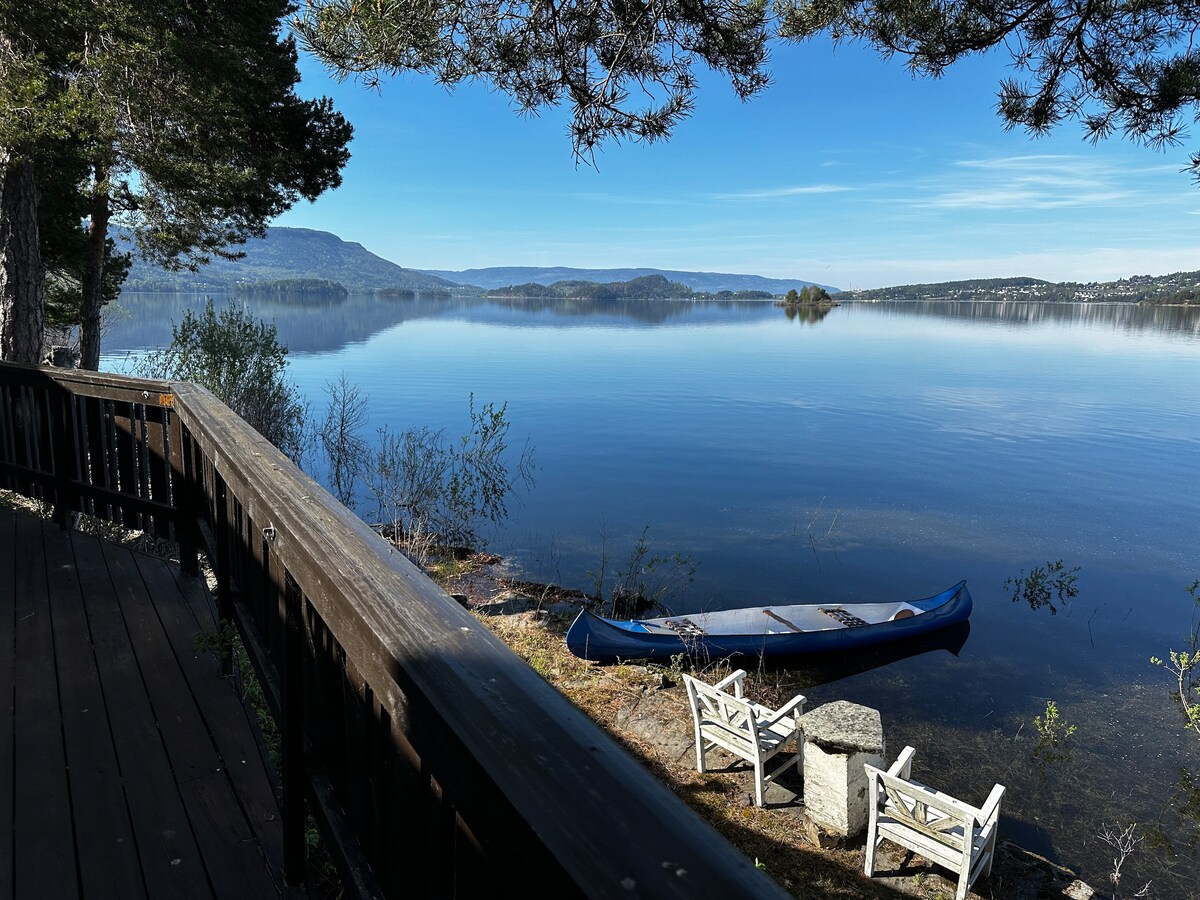 A calm water's edge is framed by trees, with a blue canoe resting on the shore. Two white chairs are positioned nearby, facing the tranquil lake. The distant mountains and clear sky create a serene backdrop, enhancing the peaceful outdoor environment.