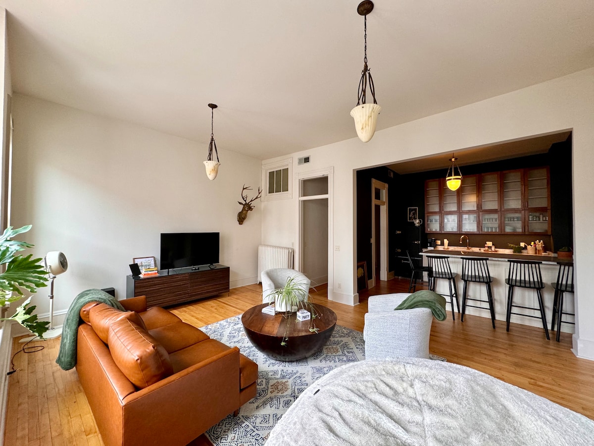 The living space features a spacious layout with natural light filtering through large windows. A comfortable leather sofa and a cozy armchair are positioned near a coffee table on a patterned area rug. An entertainment unit holds a television, and a dining area with bar stools is visible in the background.