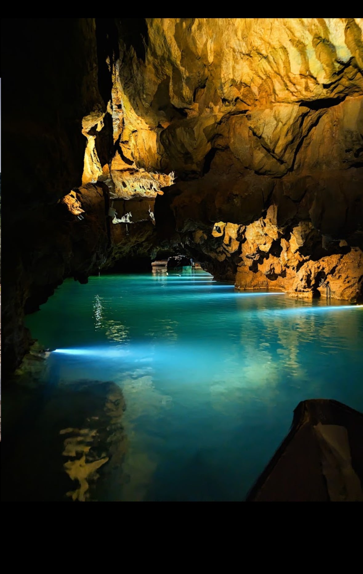 An underground cave is illuminated by soft lighting, highlighting the textured rock formations. A serene body of water reflects shades of blue, creating a calming contrast with the earthy tones of the surrounding cliffs. The tranquil atmosphere invites relaxation and exploration.