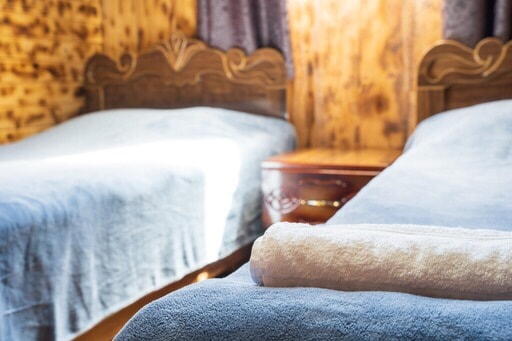 Two twin beds are dressed in soft blue blankets, positioned in a warmly lit room with wooden walls. A neatly rolled white towel is placed on one bed, and natural light filters in through the window, illuminating the space.