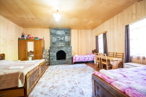 A spacious room features two wooden beds covered in soft pink linens, complemented by a cozy rug in neutral tones. A stone fireplace serves as a focal point, and a wooden dining table with chairs is positioned near large windows adorned with dark curtains.