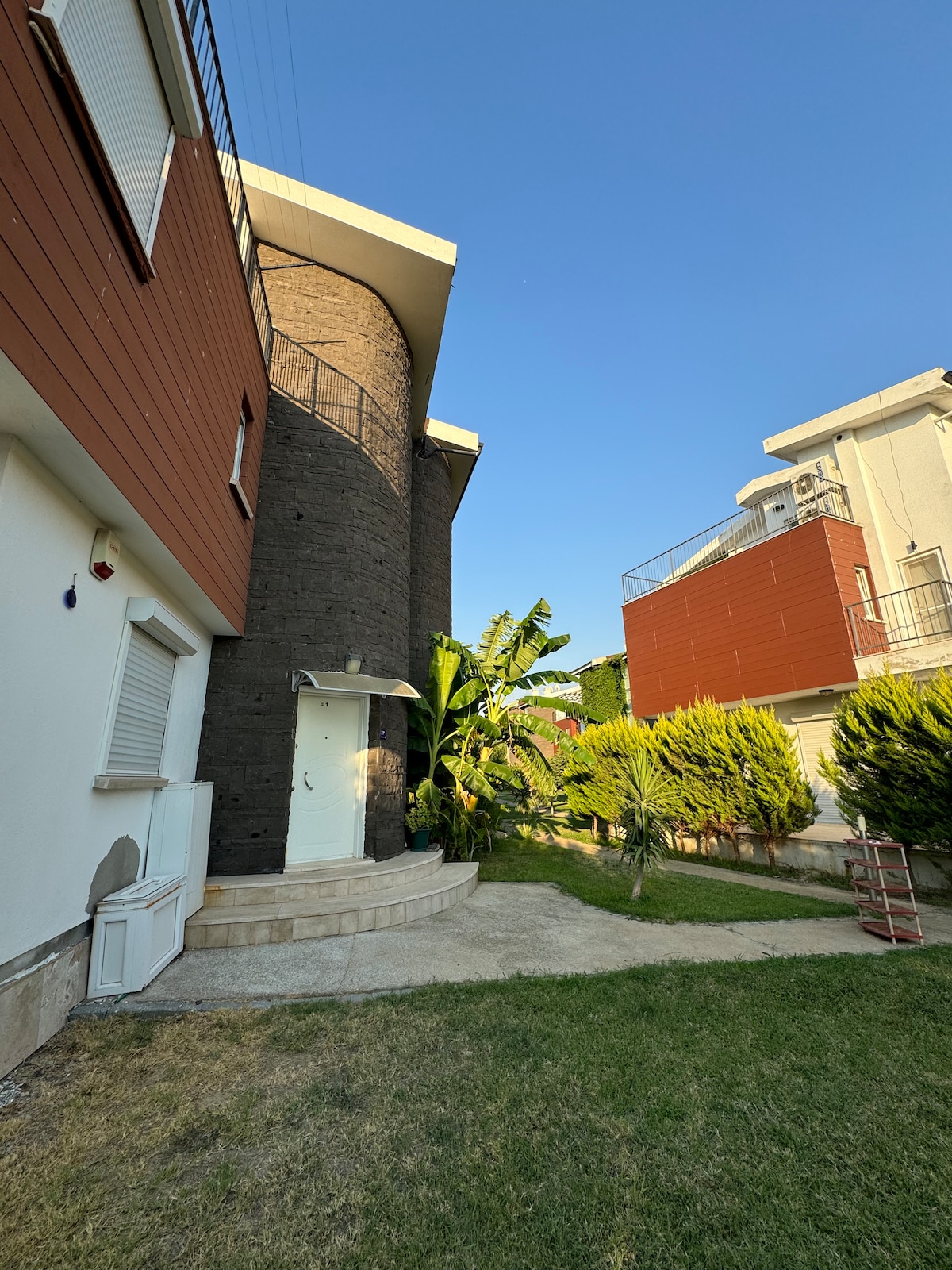 The exterior of a three-story villa is depicted, featuring a blend of stone and wood paneling. Lush green foliage surrounds the entrance steps, which lead to a welcoming door. Clear blue skies provide a bright backdrop, enhancing the villa's architectural details.