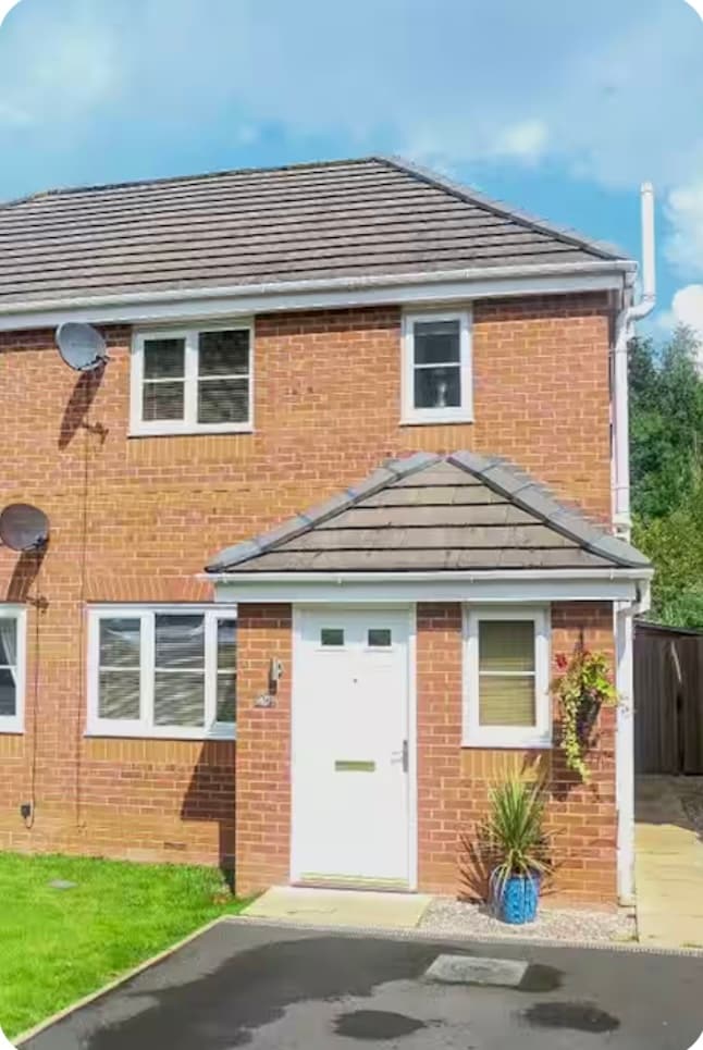 The exterior of a semi-detached house is visible, showcasing brick walls and a pitched roof. A white front door is framed by two windows, accented with a decorative potted plant beside the entrance. A small lawn and driveway complement the welcoming facade.