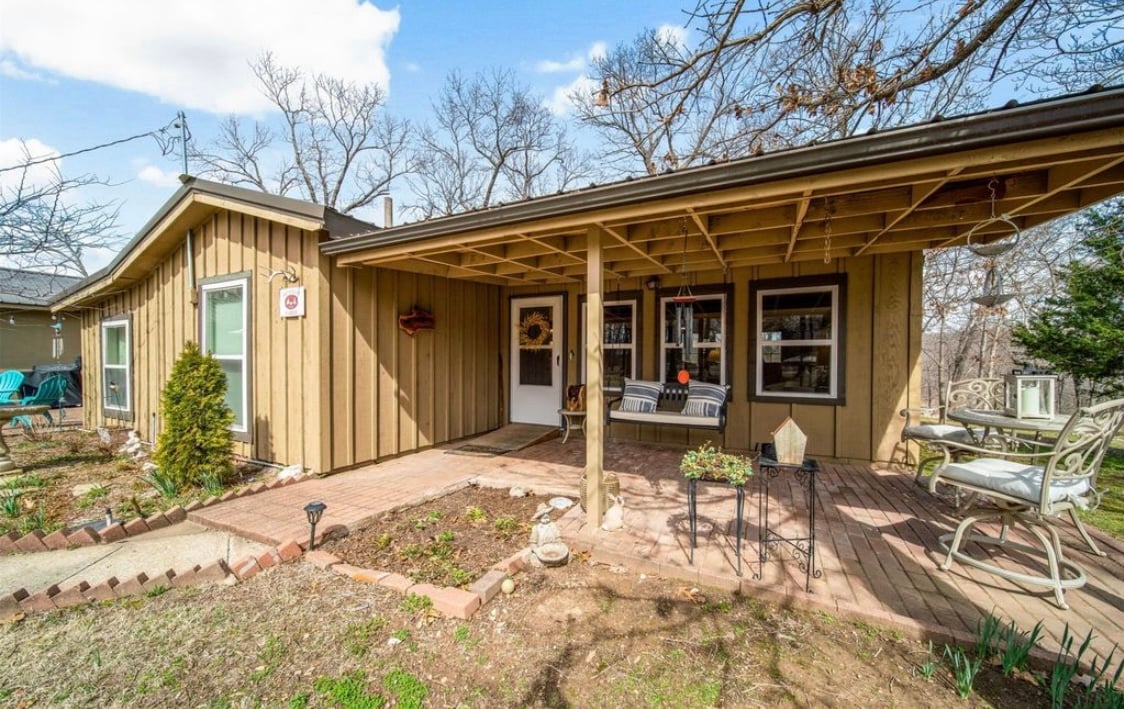 The cabin's entrance is framed by a spacious porch with a wooden roof. Outdoor seating is arranged along the porch, with a welcoming mat at the door. The landscaped yard features flower beds and a pathway leading to the entrance, surrounded by trees.