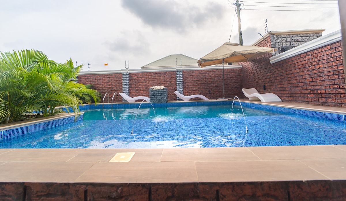 A serene outdoor pool area features vibrant blue tiles and gentle water fountains. Surrounding the pool are comfortable lounge chairs and lush greenery, enhanced by a shaded canopy. The brick wall backdrop provides a sense of privacy, creating a calm atmosphere.