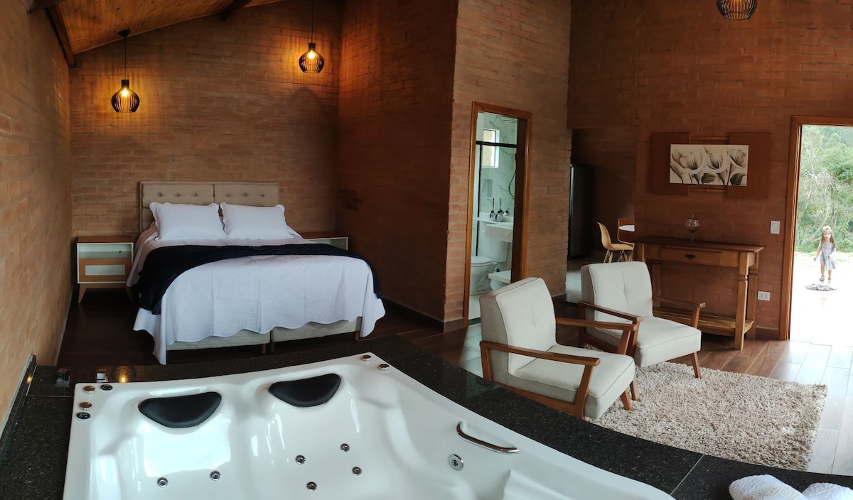 A spacious bedroom showcases a comfortable bed with soft linens, accompanied by two armchairs and a small side table. A modern hot tub is featured in the foreground, while natural light filters through large windows, highlighting the warm brick walls.